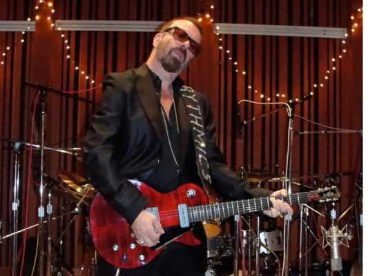 Clip-On vs Pedal Guitar Tuner: 5 Smart Tests for Loud Rooms Guitarist playing a red electric guitar beside microphones and a drum kit